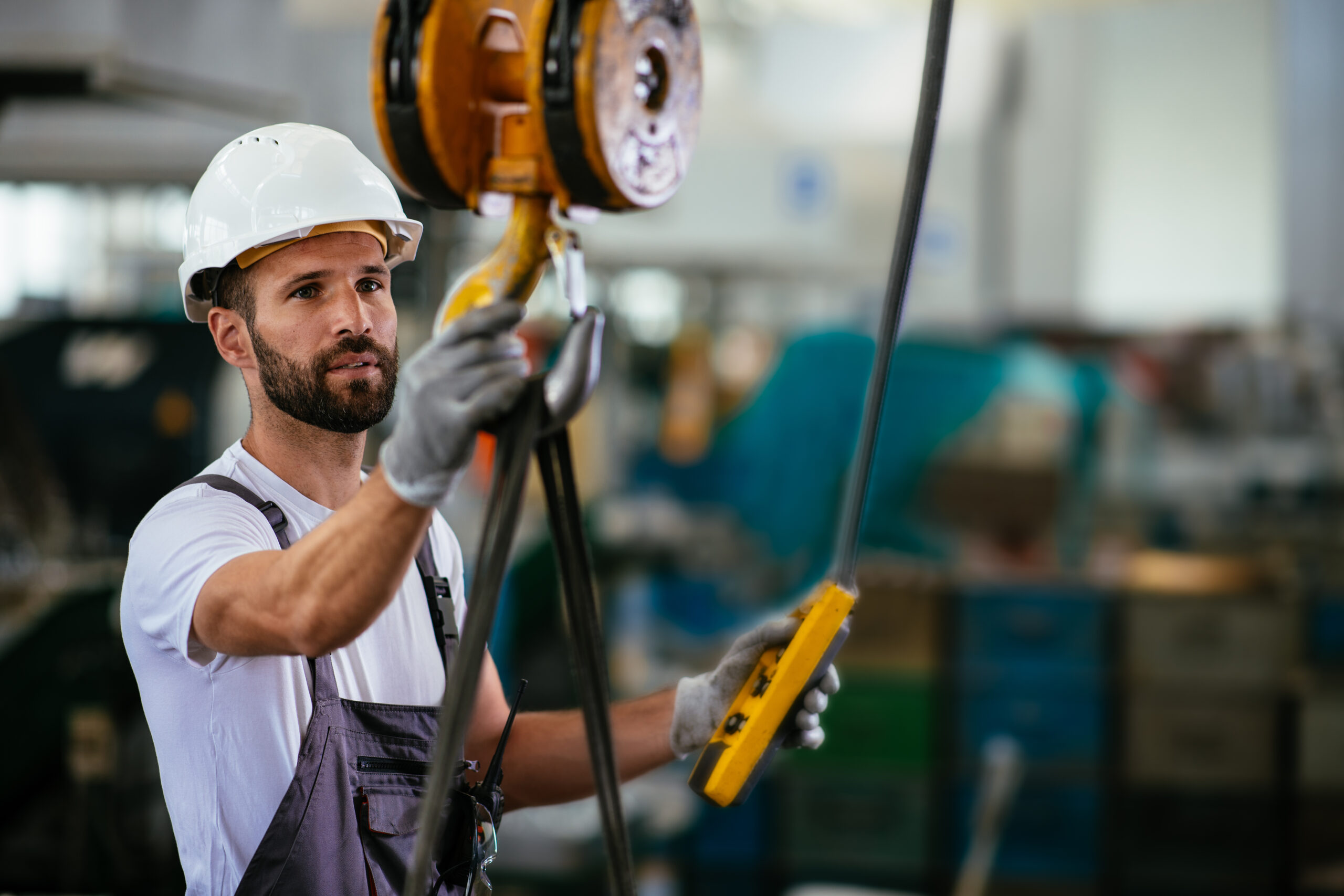 A crane operator wearing a hard hat holding a crane hook and a pendant controller in a manufacturing factory